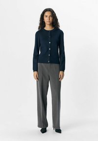 Cardigan en maille bleu marine avec col rond et fermeture à boutons sur le devant ; associé à un pantalon large gris et des chaussures à talons hauts noires.