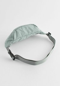 Svetlo zelena bum bag s ukrivljenim oblikom, oblazinjenim materialom in nastavljivim sivim trakom, opremljena z zaklepom za hitro odpenjanje.
