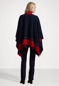 Laivastonsininen villasekoite poncho punaisilla korostuksilla, jossa on erottuva raitakuosi helmassa. Löysä istuvuus, drapeeraava siluetti ja korkea kaulus.