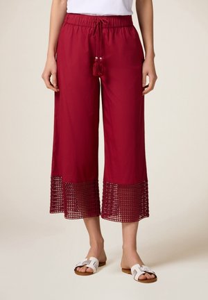Bordeauxfarbene Culottes mit elastischem Bund und Kordelzug, ausgestattet mit einem strukturierten Spitzensaum. Kombiniert mit weißen Slipper-Sandalen.