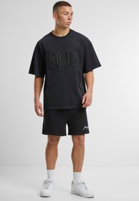 Juodas per didelis T-shirt su tekstūruota grafika, derinamas su juodais šortais, ant kurių yra baltas logo. Modelis dėvi baltus sportinius batus ir trumpas kojines.