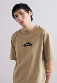 Beige bomulds T-shirt med rund hals, korte ærmer og sort grafisk tekst trykt på forsiden. Glat tekstur hele vejen igennem.