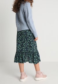 Hellblau gestrickter Pullover mit Kabelmuster, kombiniert mit einem dunklen, floralen Midirock, der grüne Blumen und einen gerafften Saum aufweist, getragen mit blassrosafarbenen Turnschuhen.