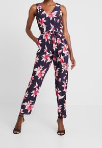 Combinaison fleurie avec un profond décolleté en V, sans manches, taille cintrée et jambes droites. Présente un fond bleu marine avec des fleurs roses et blanches.