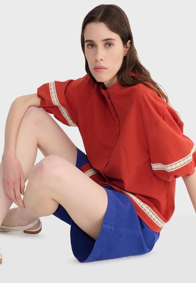 Žena s dlhými hnedými vlasmi sedí, oblečená v voľnej červenej blúzke s bielym čipkovým lemom a modrých šortkách na jednoduchom svetlom pozadí.