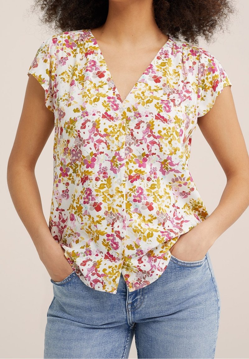 Femme portant un chemisier blanc à col en V avec des motifs floraux roses, rouges et jaunes et un jean bleu clair, mains dans les poches.