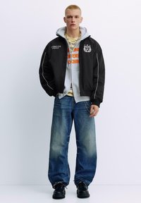 Zwarte bomberjack met witte accenten, grijze hoodie met oranje afbeelding, gestreept shirt, losse spijkerbroek en zwarte laarzen.