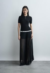 Mladá žena s dlhými tmavými vlasmi má na sebe čierne pletené polo tričko s krátkym rukávom a priehľadné čierne plisované široké nohavice na jednoduchom svetlom pozadí.