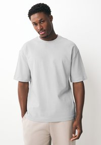 Man with short curly hair wearing a plain light grey oversized T-shirt and light beige pants, standing against a white background.