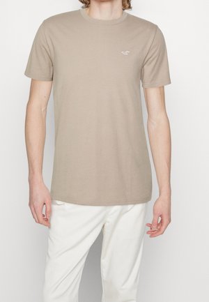 Beige katoenen t-shirt met een klassieke ronde hals, korte mouwen en een klein logo in lichtroze op de linkerborst. Rechte zoom.