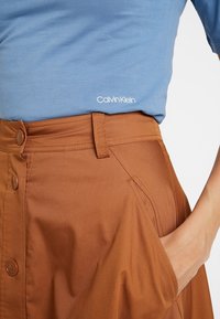 Calvin Klein vaaleansininen T-paita logolla, joka on erottuvasti housun yläpuolella ruskeassa hameessa, jossa on nappi- ja sivutaskut. Hame on valmistettu sileästä kankaasta.