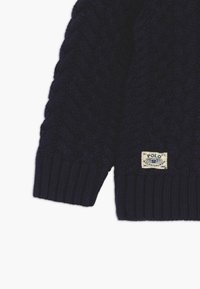 Tummansininen neulepusero, jossa on kaapelikuvio ja resorit. Oikeassa alakulmassa oleva etiketti lukee "Polo Ralph Lauren."