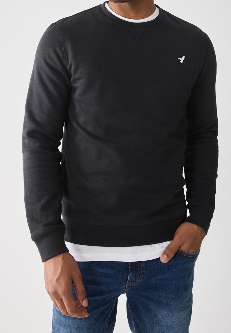 Mężczyzna w czarnej bluzie z okrągłym dekoltem z białym logo, białej podkoszulce i niebieskich dżinsach, stoi z jedną ręką w kieszeni.