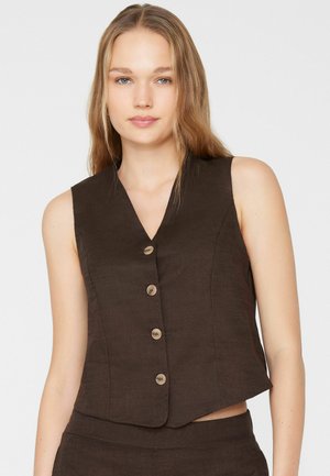 Vrouw met lang blond haar, gekleed in een mouwloos donkerbruin vest met knopen en bijpassende broek, staand tegen een effen lichte achtergrond.