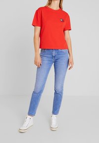 Maglietta rossa in cotone con scollatura rotonda e maniche corte, caratterizzata da un piccolo logo ricamato sul petto. Abbinata a jeans azzurri chiari e scarpe da ginnastica bianche.