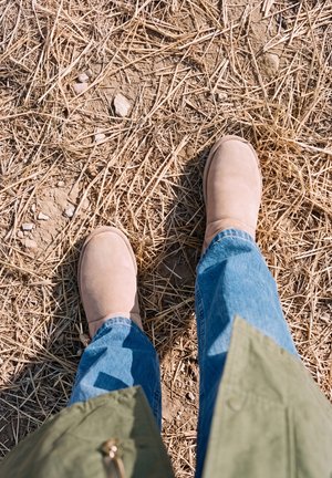 Une personne portant des bottes beiges et un jean bleu se tient sur de la paille sèche et de la terre, vue d’en haut.