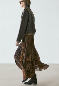 Bruine leren jas over een patroonrijke, vloeiende, gelaagde rok in aardetinten, gecombineerd met zwarte enkellaarzen en donkere zonnebril.