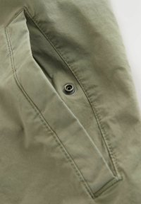 Nahaufnahme einer olivgrünen Stofftasche mit einem Metall-Druckknopf und sichtbaren Nähten.