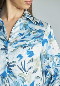 Camisa de botones con un patrón floral en tonos azules y verdes, hecha de una tela suave con collar clásico y botones parecidos a perlas.