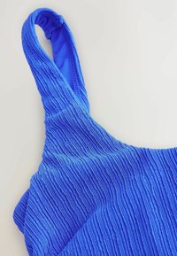 Débardeur en tissu bleu avec un motif texturé et côtelé. Présente des bretelles ajustables et une doublure intérieure lisse.
