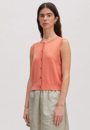 Vrouw met lang donker haar, gouden oorringen, een mouwloos koraalkleurig topje met knopen en een lichtbeige rok met hoge taille, staand tegen een witte achtergrond.