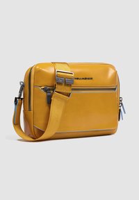 Borsa gialla in pelle con chiusura a zip, tracolla regolabile, tasca frontale e accenti blu. Presenta una texture liscia e una forma rettangolare.