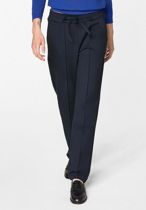 Persoon die zwarte, op maat gemaakte broek met een trekkoord in de taille, een blauw shirt met lange mouwen en zwarte loafers draagt, lopend naar voren.