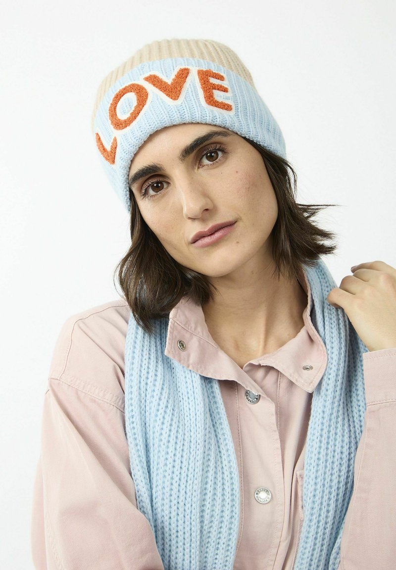 Gebreide beanie in lichtblauw met een crèmekleurige top, met daarop "LOVE" in oranje. Gepaard met een textuur van een lichtblauwe sjaal en een roze shirt.
