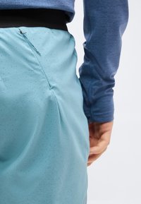 Άτομο που φοράει φούστα ανοιχτού μπλε χρώματος με μικρές βούλες και φερμουάρ στο πλάι, σε συνδυασμό με μπλε μπλούζα μακριάς μανσέτας, με το χέρι χαλαρωμένο δίπλα στο σώμα.