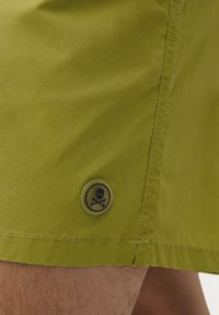 Shorts verts en tissu léger, arborant un patch arrondi avec un emblème de skull et os croisés sur le bas à gauche.