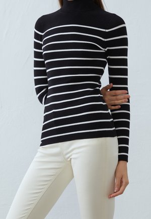 Frau trägt einen schwarz-weiß gestreiften Rollkragenpullover mit cremefarbenen Hosen und posiert mit einem verschränkten Arm.