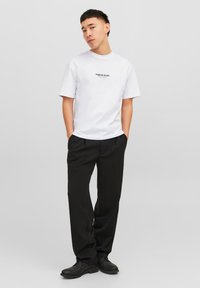 White cotton t-shirt with short sleeves featuring a black "Originals Studio" logo. Paired with black trousers and black shoes.