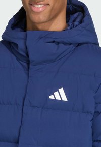 Tmavomodrá prešívaná bunda s vysokým golierom a kapucňou; na hrudi je biely logo. Materiál pôsobí hladko s prešivanými sekciami.