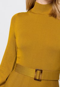 Senapsgul ribbad polotröja med långa ärmar och ett brett matchande bälte som har en rektangulär brun spänne, detaljerad textur.