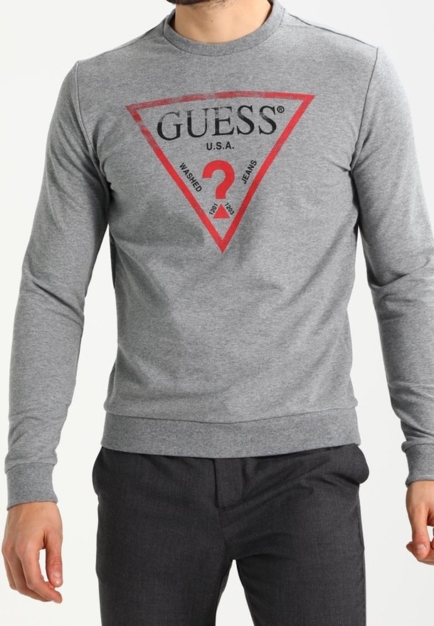 Hombre vestido con una camisa de manga larga gris con un logo triangular rojo de GUESS y pantalones negros, cortados por debajo de la barbilla.