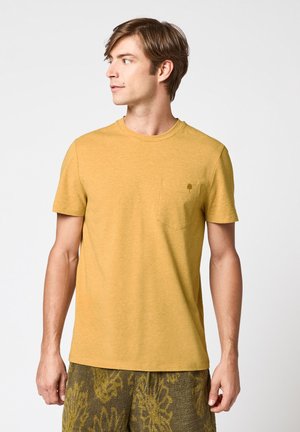 Jeune homme regardant vers la gauche, portant un t-shirt jaune moutarde avec une poche poitrine et un short vert à motif, sur un fond uni.