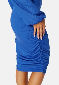 Abito blu a coste con dettagli arricciati ai lati, maniche lunghe e silhouette aderente, che mette in evidenza un tessuto liscio e strutturato.