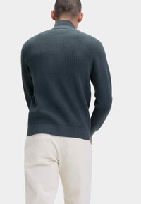 Dunkelblauer, gerippter Pullover mit hohem Kragen, langen Ärmeln und taillierter Taille. Kombiniert mit hellen Hosen, die einen strukturierten Stoff zeigen.