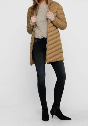 Mujer con un abrigo acolchado beige sobre un suéter de punto beige, vaqueros pitillo negros y botines negros de tacón con punta afilada.