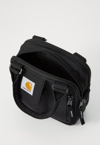 Sac en tissu noir avec double fermeture éclair, poignées supérieures et une badge logo blanc. Design compact avec une surface texturée.