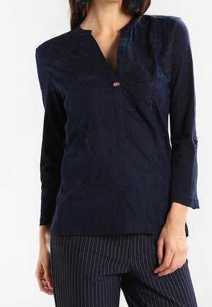 Kobieta ubrana w granatową bluzkę z długim rękawem z delikatnym kwiatowym wzorem i małym detalem flagi przy dekolcie, w połączeniu z spodniami w prążki.