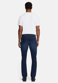 Jeans blu scuro con vestibilità slim, realizzati in denim. La vista posteriore mostra due tasche posteriori e un elastico con passanti per cintura. Abbinati a una t-shirt bianca.