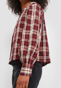 Femme portant une chemise à carreaux rouge, blanche et noire à manches longues avec des manches bouffantes et des poignets boutonnés, associée à un jean noir.