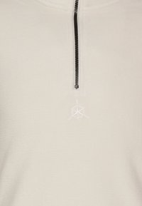 Maglione in pile color crema con una cerniera nera e un piccolo logo a forma di stella dei marinai ricamato sotto la cerniera.