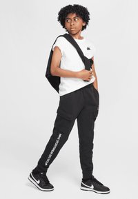 Pantalones cargo negros con texto blanco, combinados con una camiseta blanca de manga corta. Zapatillas negras y una bolsa negra colgada del hombro.