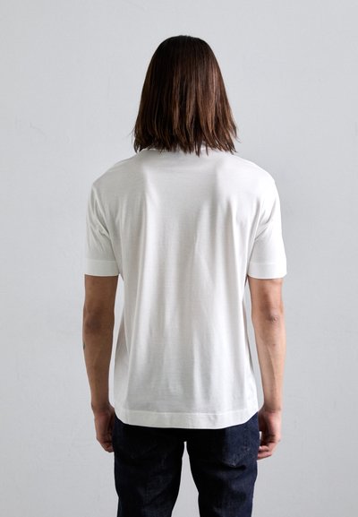 Person with shoulder-length brown hair wearing a plain white short-sleeve shirt and dark pants, standing against a light gray background.