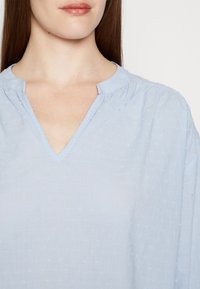 Blusa azul clara com decote em V, corte descontraído e padrão de bolinhas texturizadas. O material parece suave e leve.