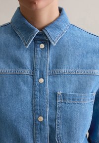 Blue denim shirt with a pointed collar, front pocket, and silver snap buttons. Features visible stitching and a smooth, textured fabric.