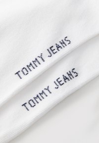 Fehér zoknik, amelyekre a "TOMMY JEANS" felirat van nyomtatva tengerészkék színben, tiszta és sima pamut textúrában.