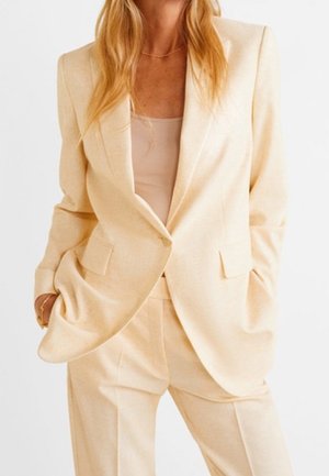 Blonde Frau trägt einen hellbeigen Anzug mit Blazer und passenden Hosen, Hände in den Taschen und darunter ein hautfarbener Top.